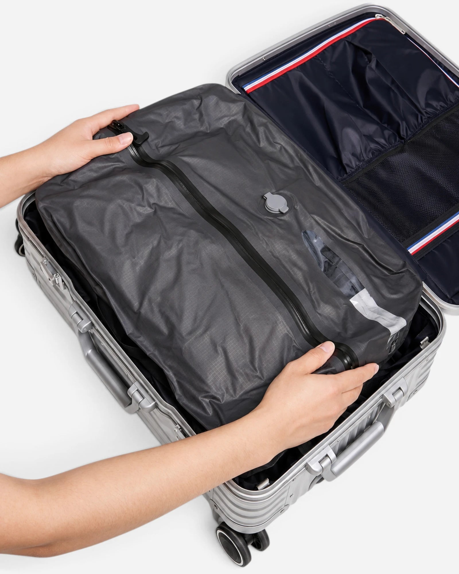 Hands placing Lavnex Rilvac™ Jet Black vacuum compression bag inside open silver hard‑shell suitcase with dark lining and mesh divider